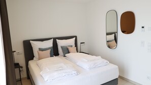Un letto rifatto con lenzuola e cuscini bianchi, un comodino con una lampada e uno specchio fissato alla parete.