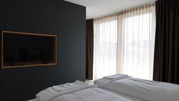 A hotel room with a large window, a flat-screen TV mounted on the wall, and two beds with white bedding.