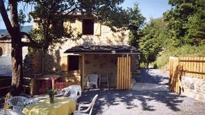 A rustic stone building with a covered patio, outdoor seating, and a gravel path leading to a wooden gate.