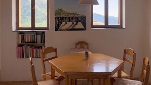 A dining area with a wooden table and chairs, two windows with mountain views, a bookshelf, and a pendant light.