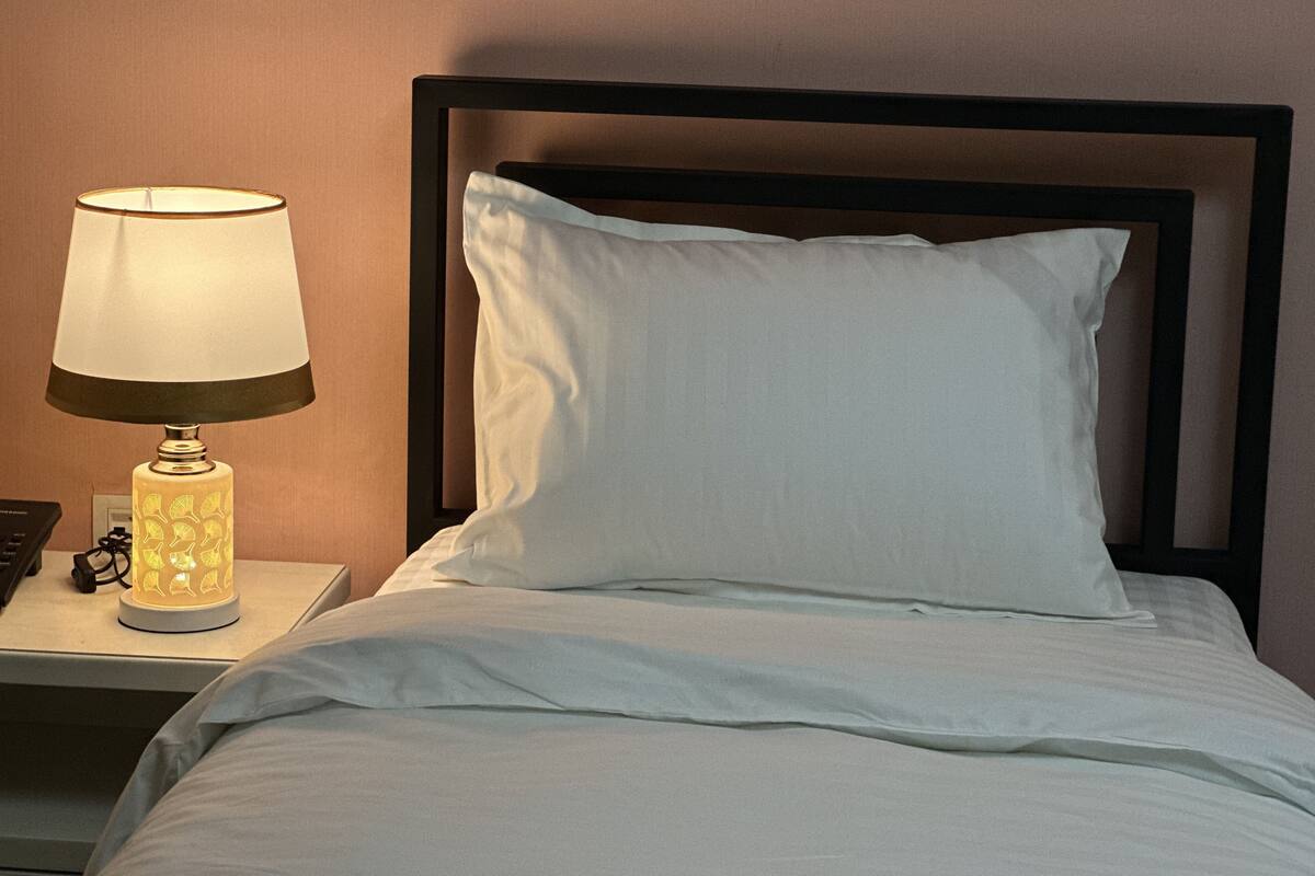 Ein ordentlich bezogenes Bett mit weißen Leinen, einem schwarzen Kopfteil und einer Nachttischlampe mit weißem Lampenschirm.