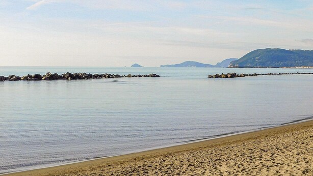 Una playa con aguas tranquilas, un espigón rocoso y colinas a lo lejos.
