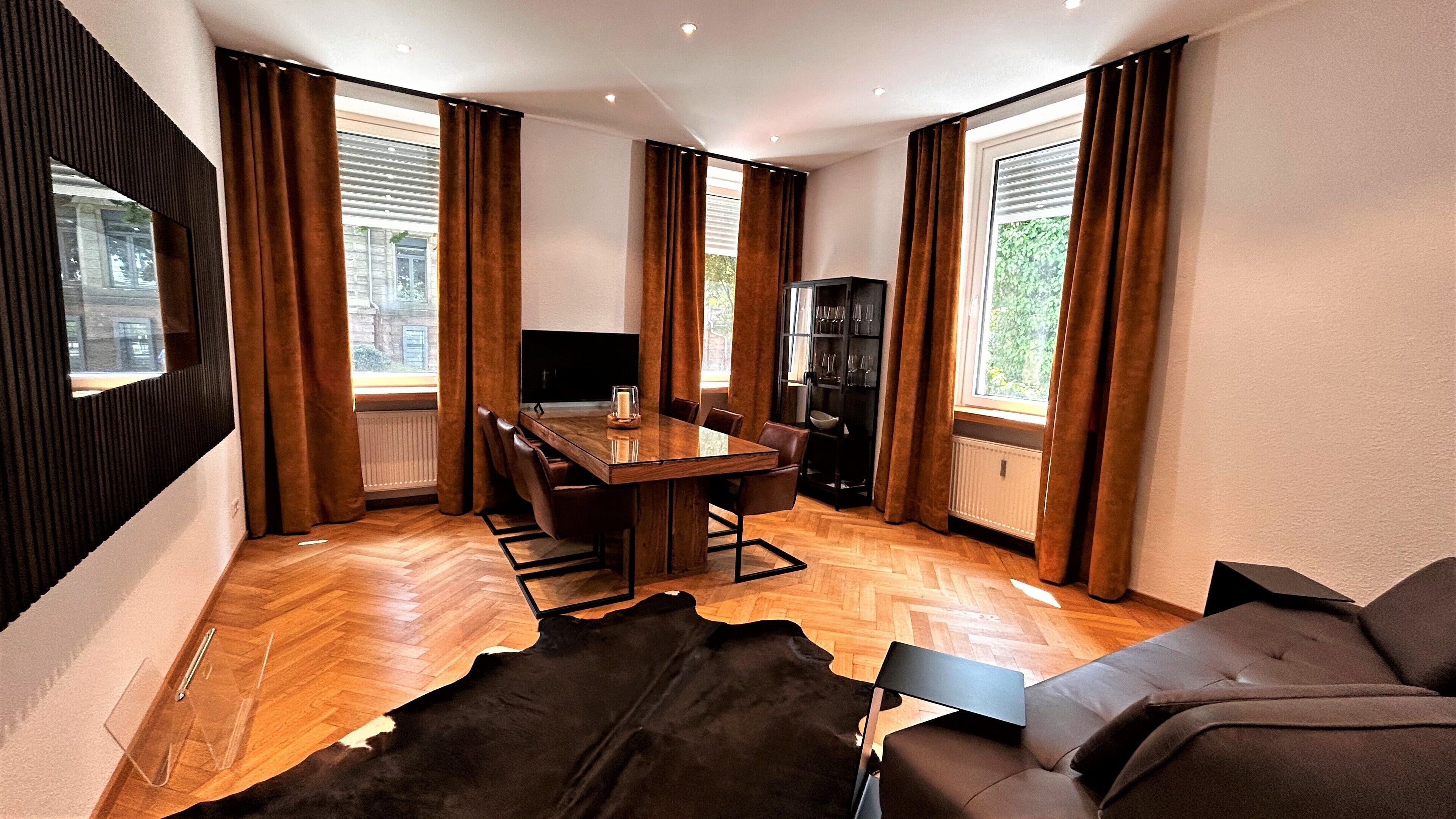 A modern living room with a wooden herringbone floor, a leather sofa, a glass-top dining table with chairs, a black entertainment unit, and large windows with brown curtains.