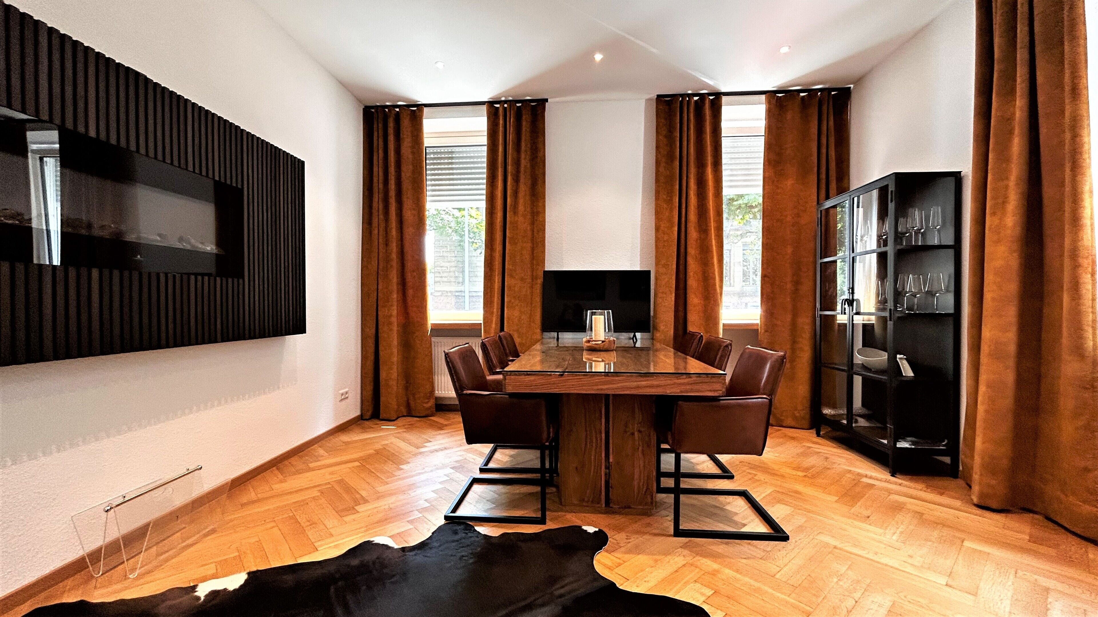 A modern dining area with a wooden table, leather chairs, a mounted TV, and a glass cabinet with wine glasses.