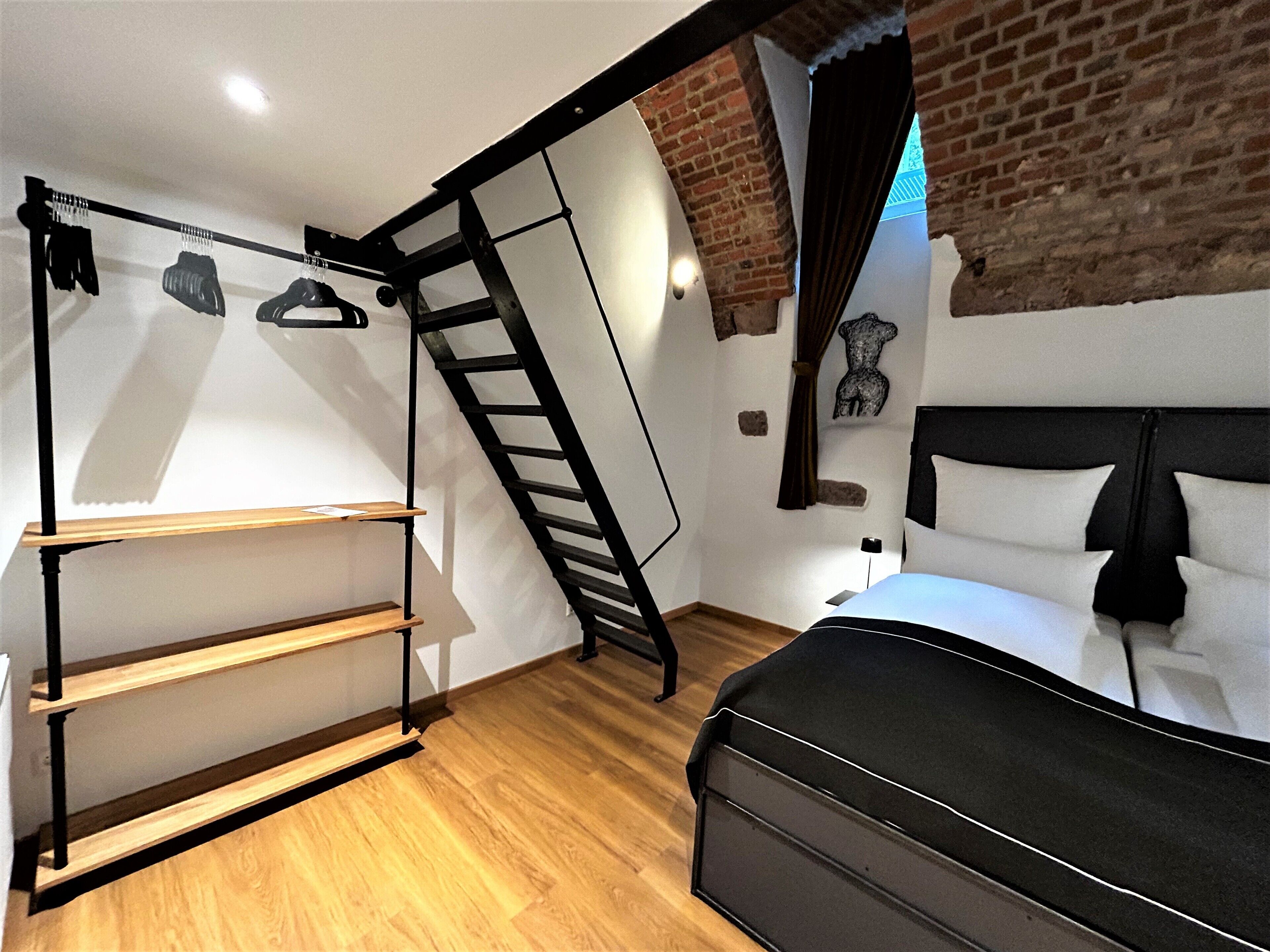 A bedroom with a loft bed, a wooden shelf unit, a ladder, and a brick wall.