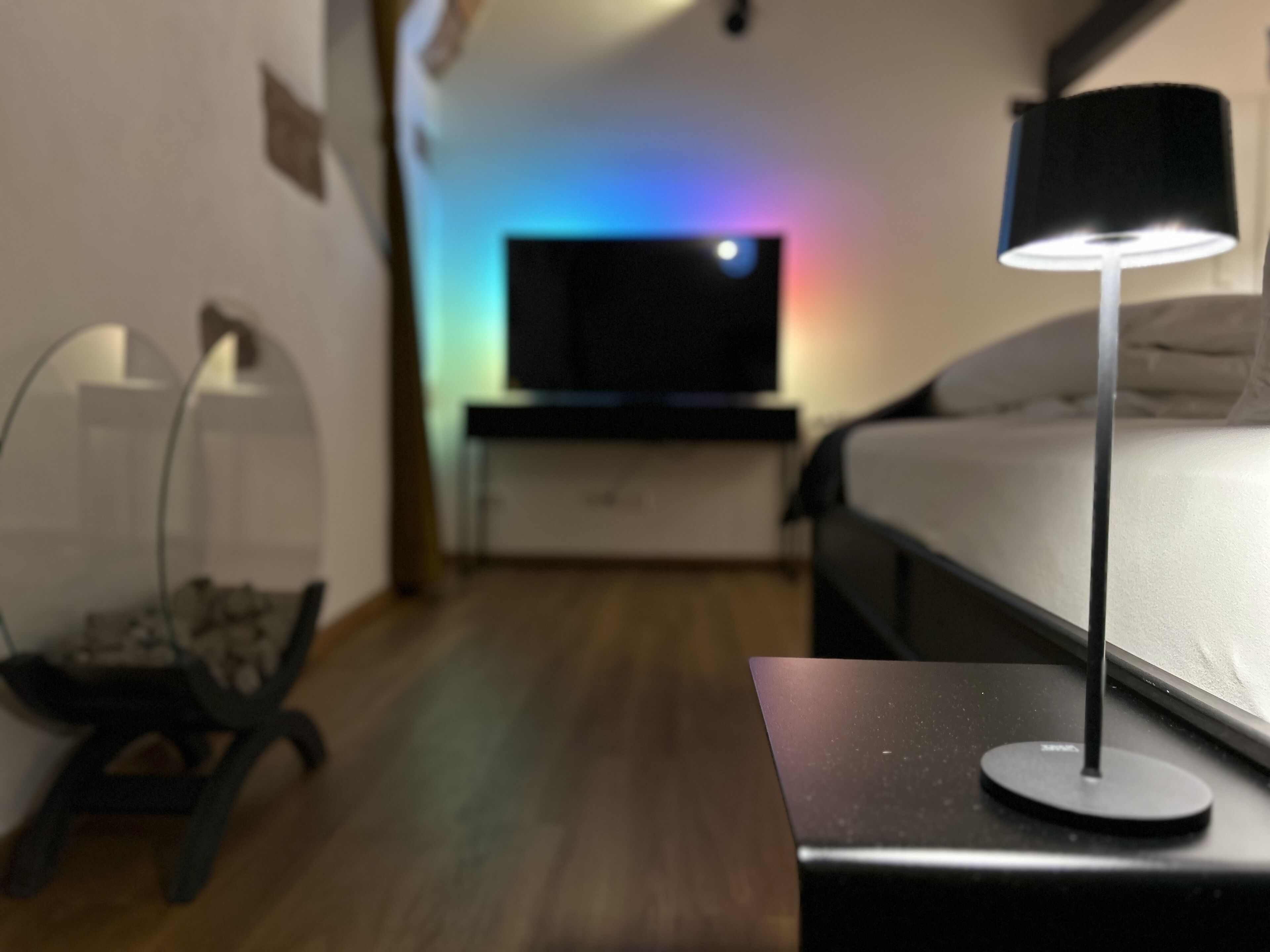 Una habitación de hotel moderna con cama, una mesita de noche con lámpara, televisión y un pequeño rincón de descanso con una silla y una mesa.