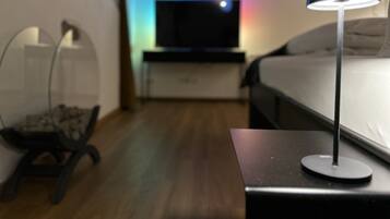 Una habitación de hotel moderna con cama, una mesita de noche con lámpara, televisión y un pequeño rincón de descanso con una silla y una mesa.