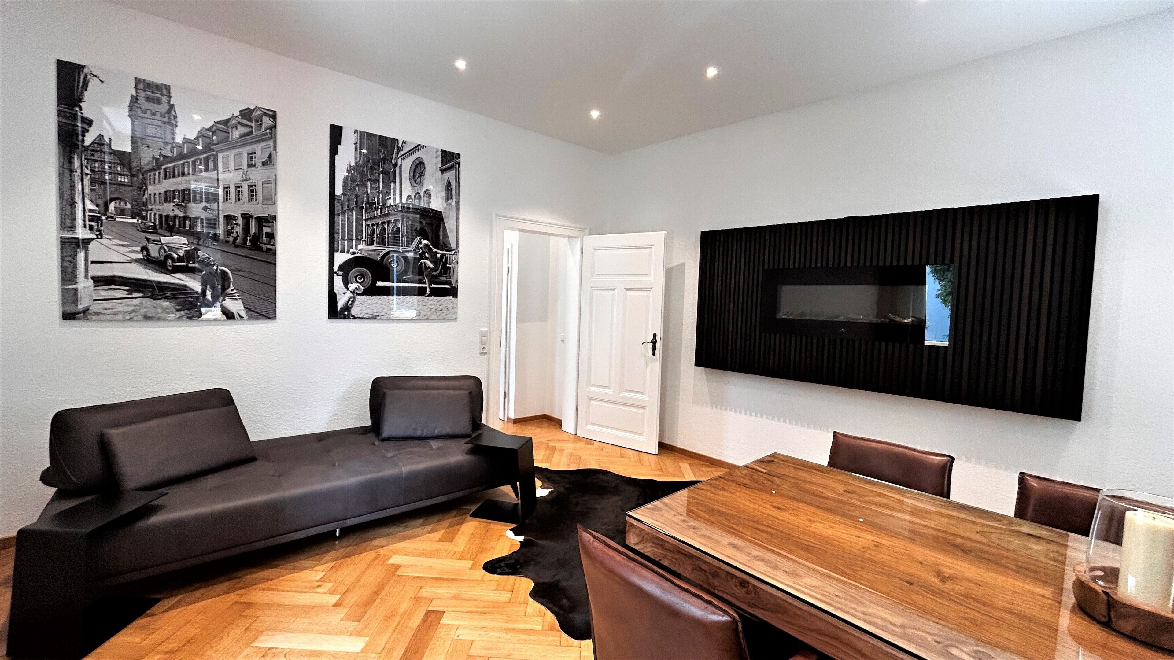 A modern living room with a sofa, a dining table, and a mounted TV. There are two black and white photographs on the wall.