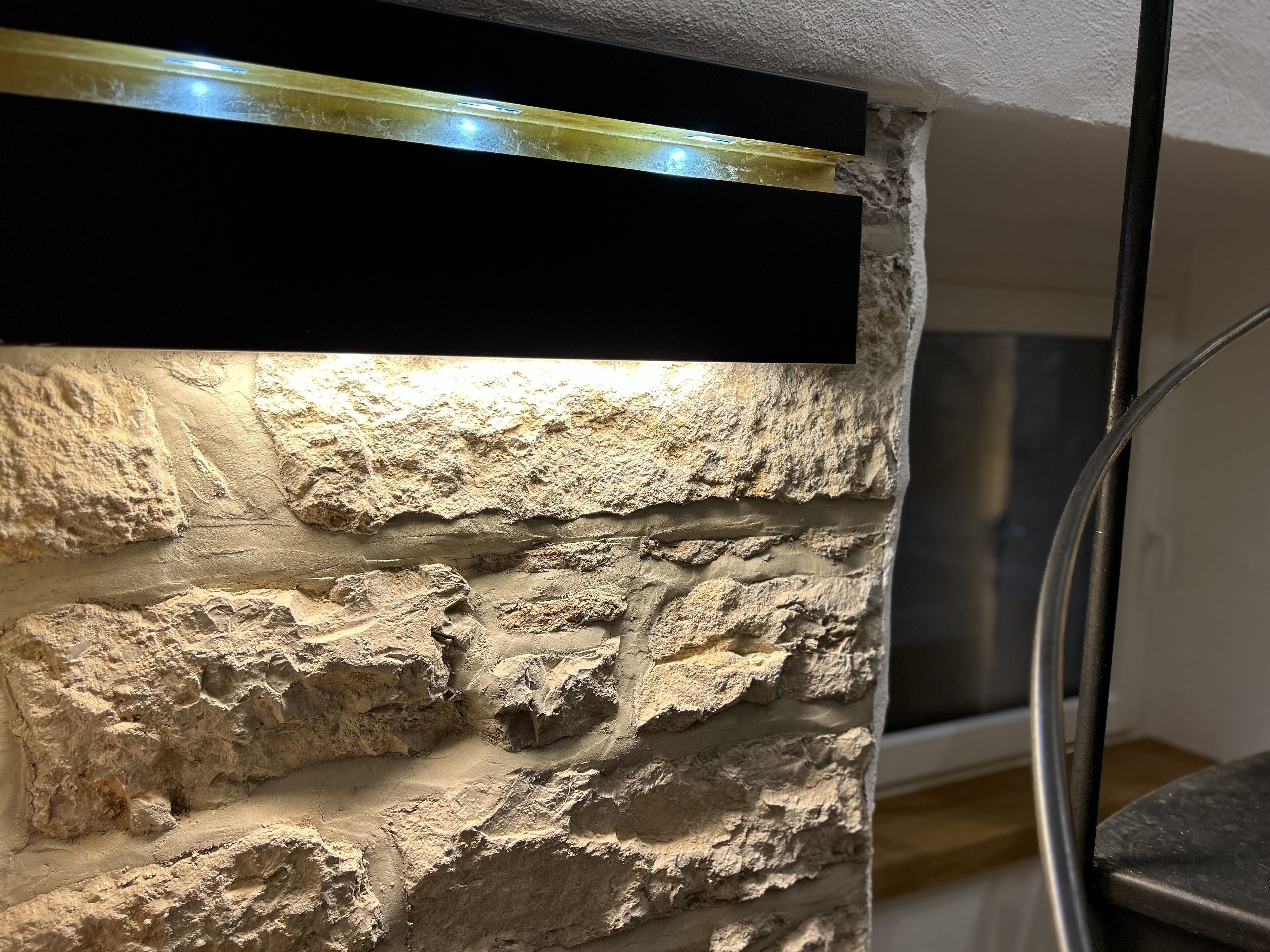 A textured stone wall with a black horizontal element, possibly a shelf or panel, and a metal structure to the right.