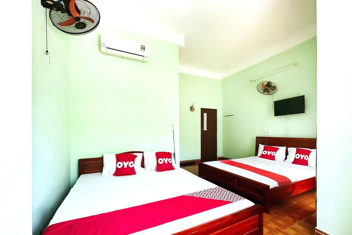 A hotel room with two beds, each with a red and white bedspread and pillows with 'OYO' branding, a wall-mounted air conditioner, a ceiling fan, a television, and a door.