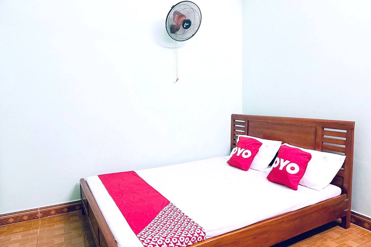 A neatly made bed with a red and white patterned bedspread, two red pillows with the word "YOYO" on them, and a wall-mounted fan.