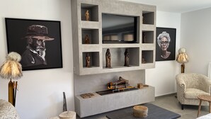 A modern living room with a grey accent wall, a flat-screen TV, and wooden sculptures. There is a fireplace with logs and a round wooden table.