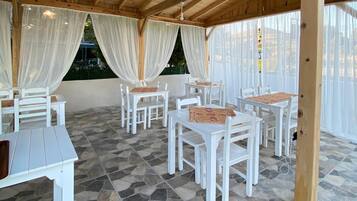 Un espacio de estar al aire libre cubierto con mesas y sillas blancas, suelo de piedra y cortinas blancas translúcidas.