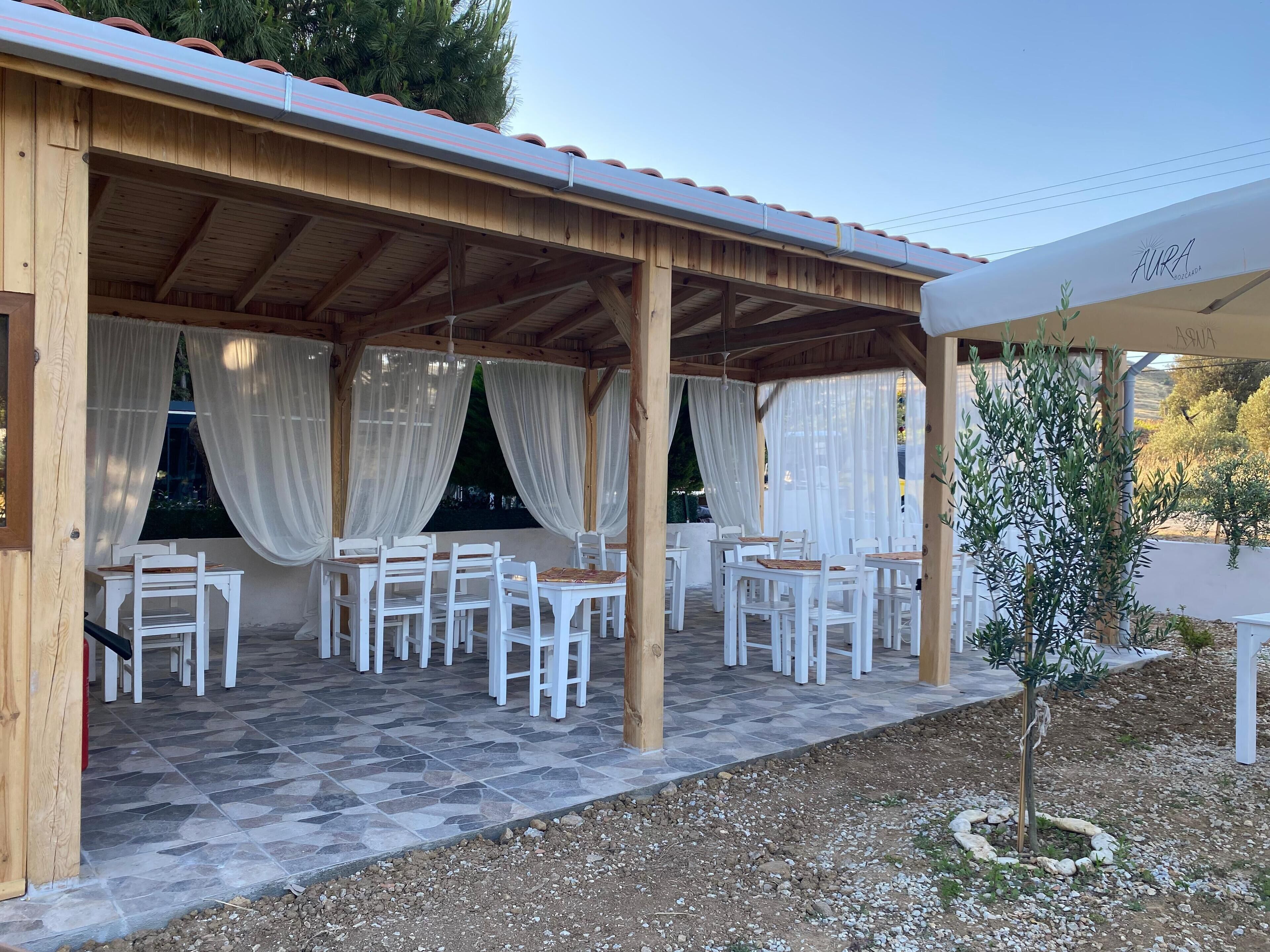 A covered outdoor seating area with white chairs and tables, a wooden pergola, and a view of the surrounding landscape.