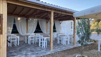 A covered outdoor seating area with white chairs and tables, a wooden pergola, and a view of the surrounding landscape.