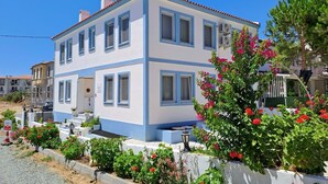 A two-story building with a blue and white exterior, multiple windows, and a small garden with red flowers. - Aura Bozcaada (Bozcaada)