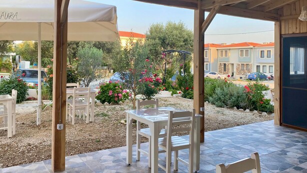 An outdoor patio area with wooden tables and chairs, a covered seating area, and a view of a residential neighborhood. - Aura Bozcaada (Bozcaada)