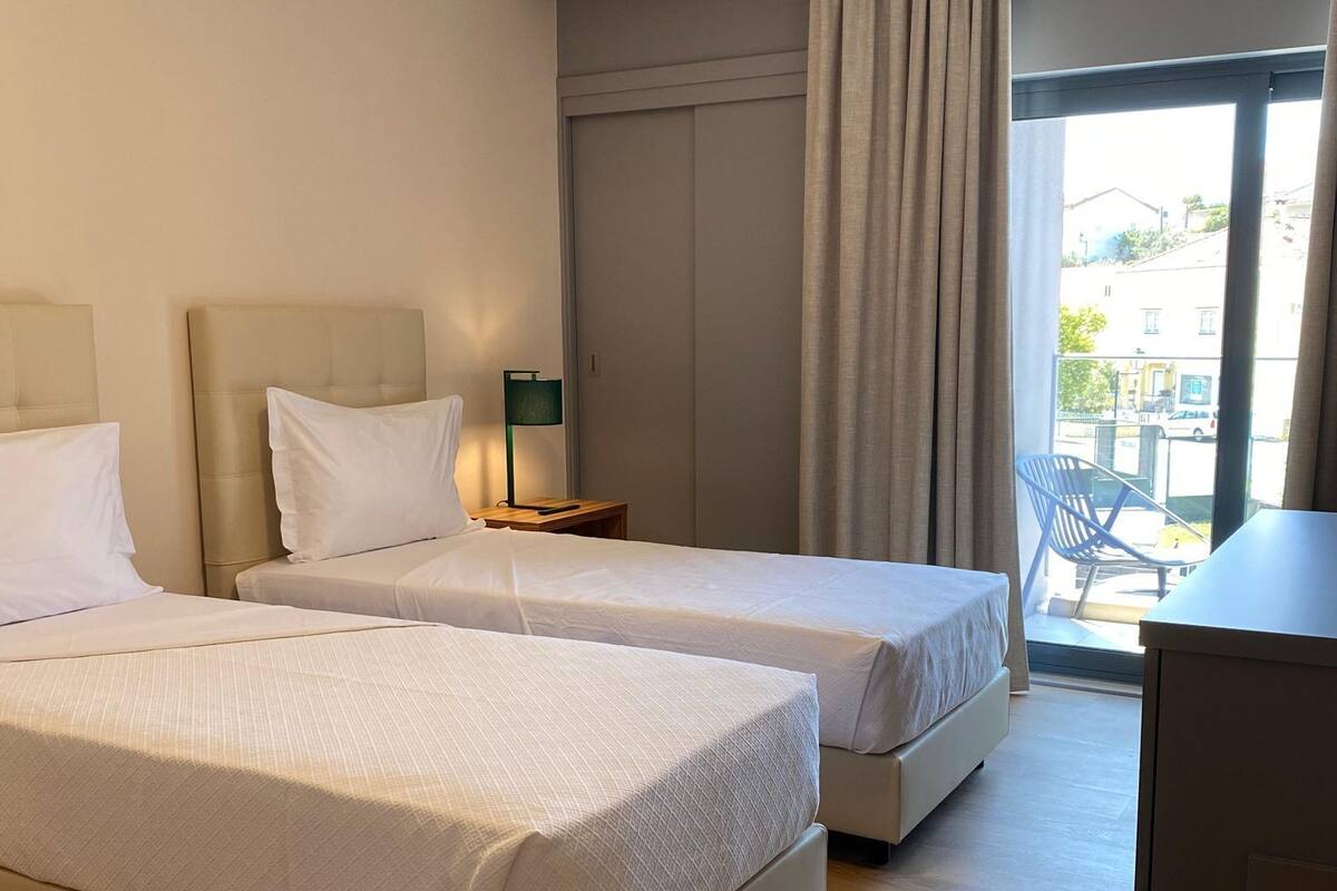 A hotel room with two beds, a desk, a chair, and a view of the outside.