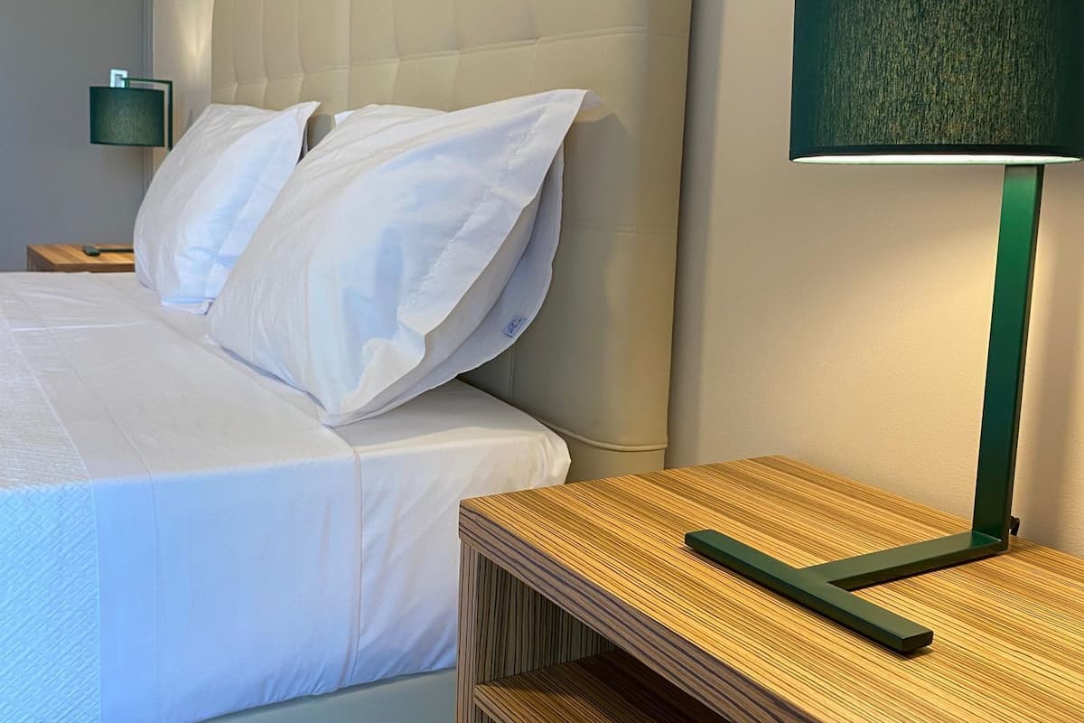 A neatly made bed with white linens, a green lamp on a wooden nightstand, and a wall-mounted lamp.