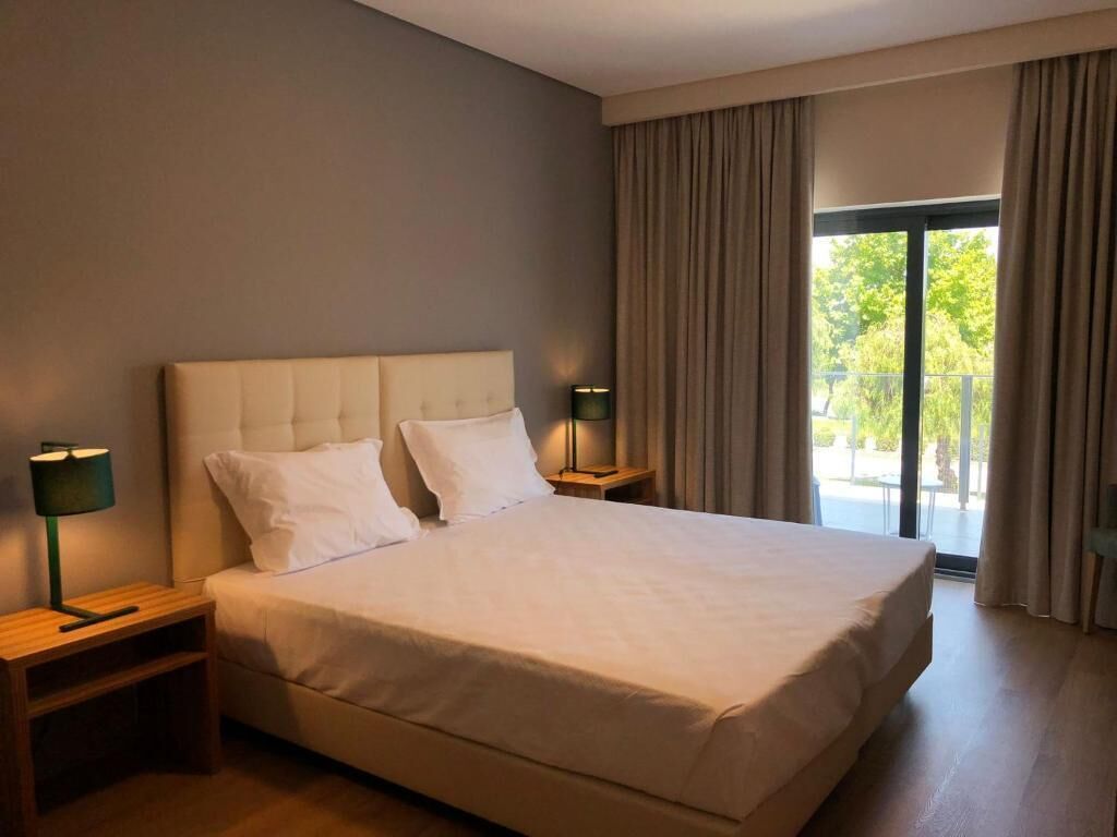 A large bed with a tufted headboard, two bedside tables with lamps, and a view of the outdoors through a large window.