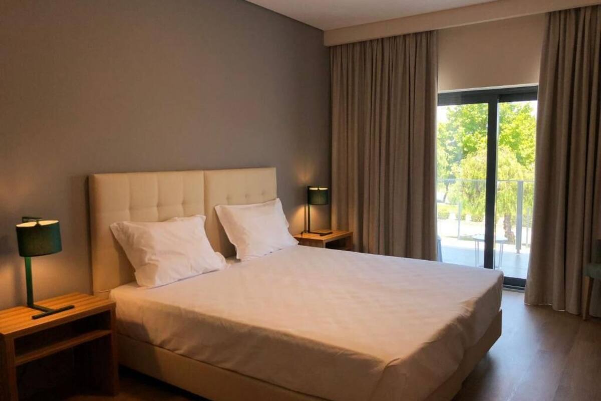 A large bed with a tufted headboard, two bedside tables with lamps, and a view of the outdoors through a large window.