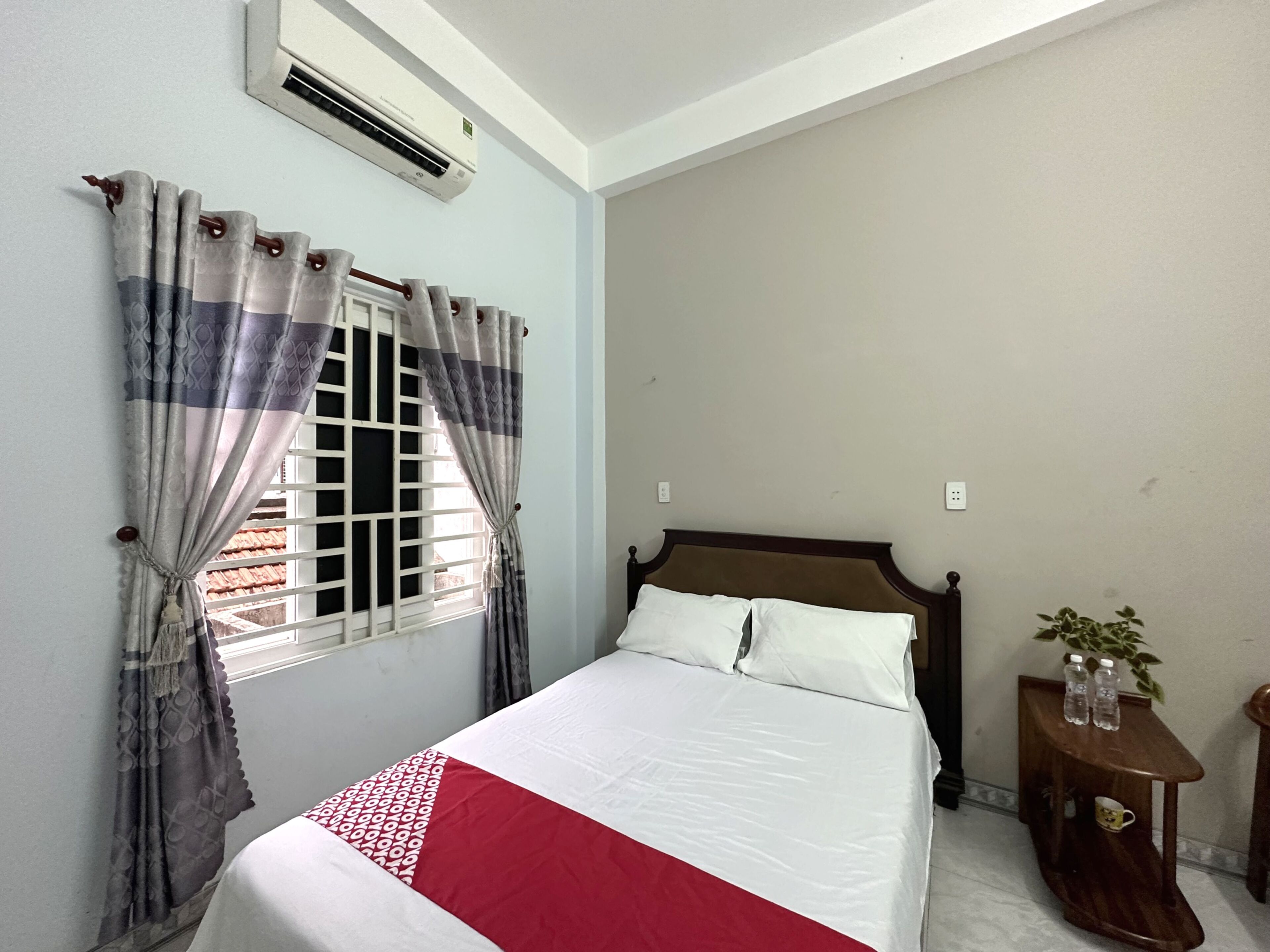 Un dormitorio con cama, ventana con cortinas, unidad de aire acondicionado, mesita de noche con planta y toma de corriente.