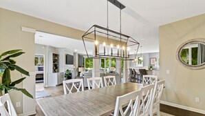 Un vaste espace de salle à manger avec une table en bois et des chaises blanches, un luminaire suspendu moderne et une vue sur l’espace de vie adjacent.