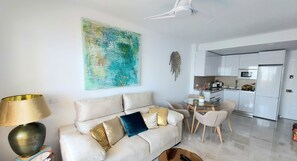 A modern living room with a beige sofa, a glass coffee table, and a dining area with a small table and chairs. A kitchen with white cabinets and appliances is visible in the background. - Benal Beach. Beautiful penthouse in first line beach.4 people. 5 swimming pools. (Benalmadena)