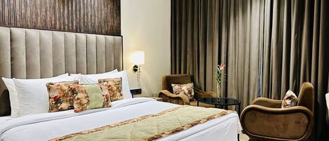 A hotel room with a large bed, a headboard, a seating area with two chairs, a small table, and a curtain backdrop.