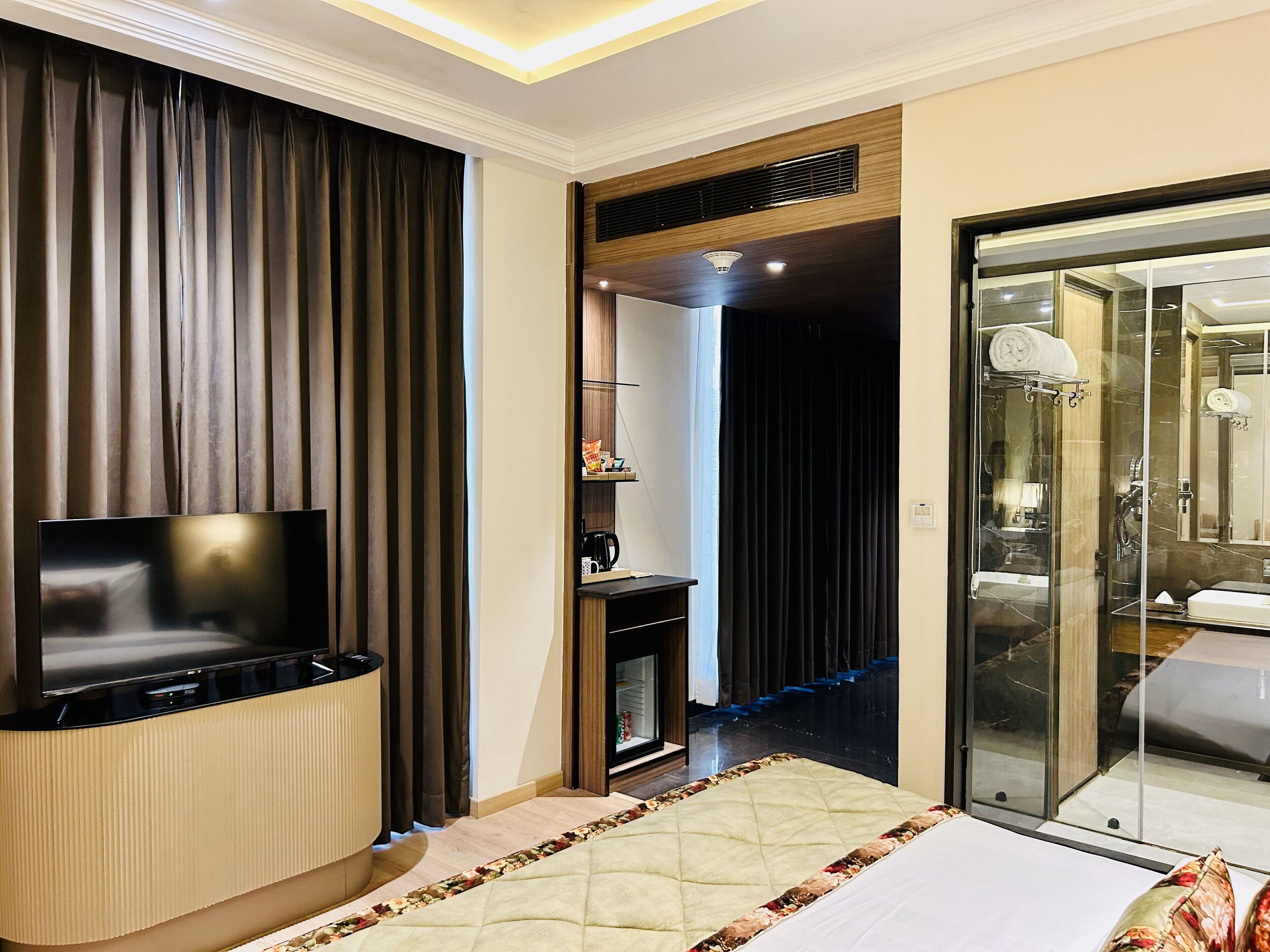 Ein Hotelzimmer mit Flachbildfernseher, einem Bett mit blumigem Überwurf, einer gläsernen Duschkabine und einem dunklen Vorhang.