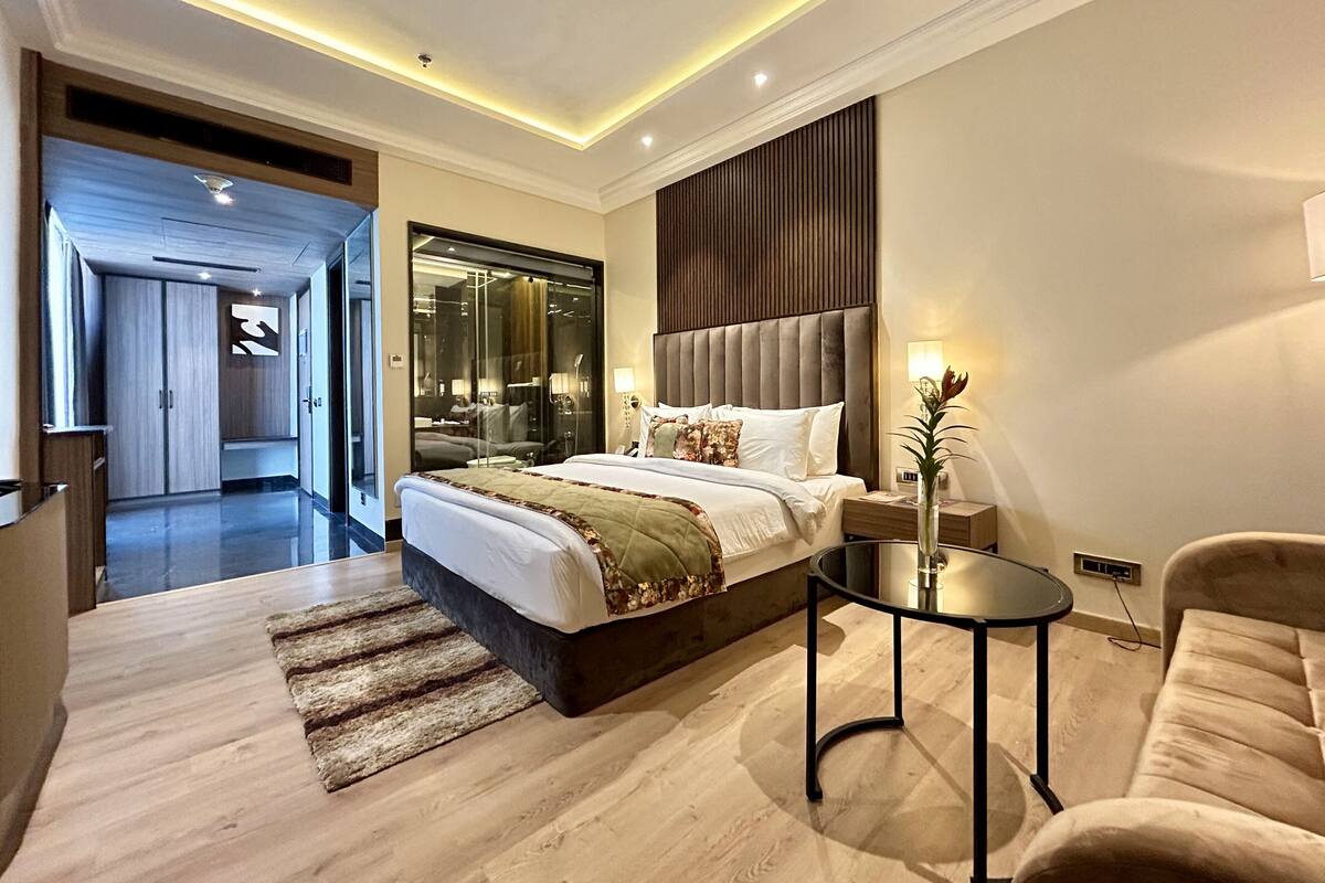 A modern hotel room with a large bed, a sitting area with a sofa and a small table, and a separate bathroom visible through a glass door.