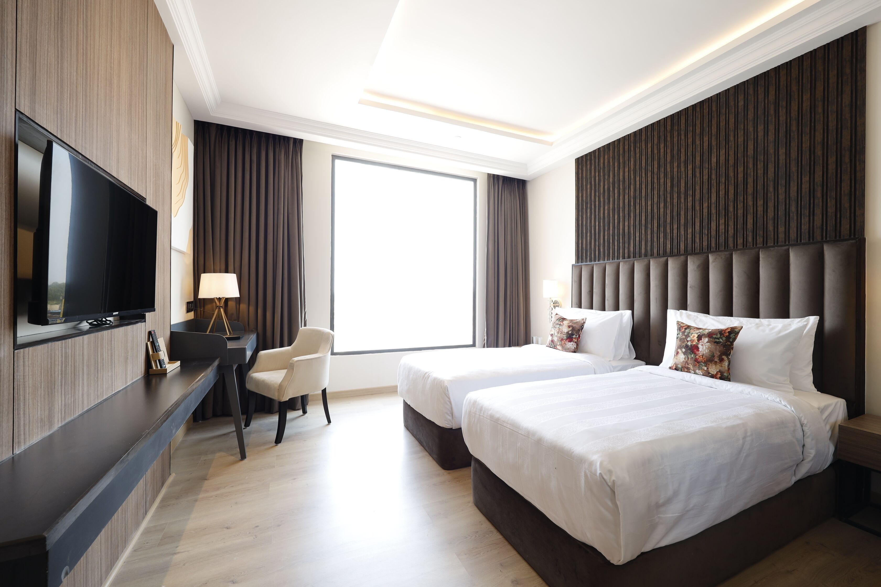 A hotel room with a flat-screen TV, two beds, a desk with a chair, and a large window.