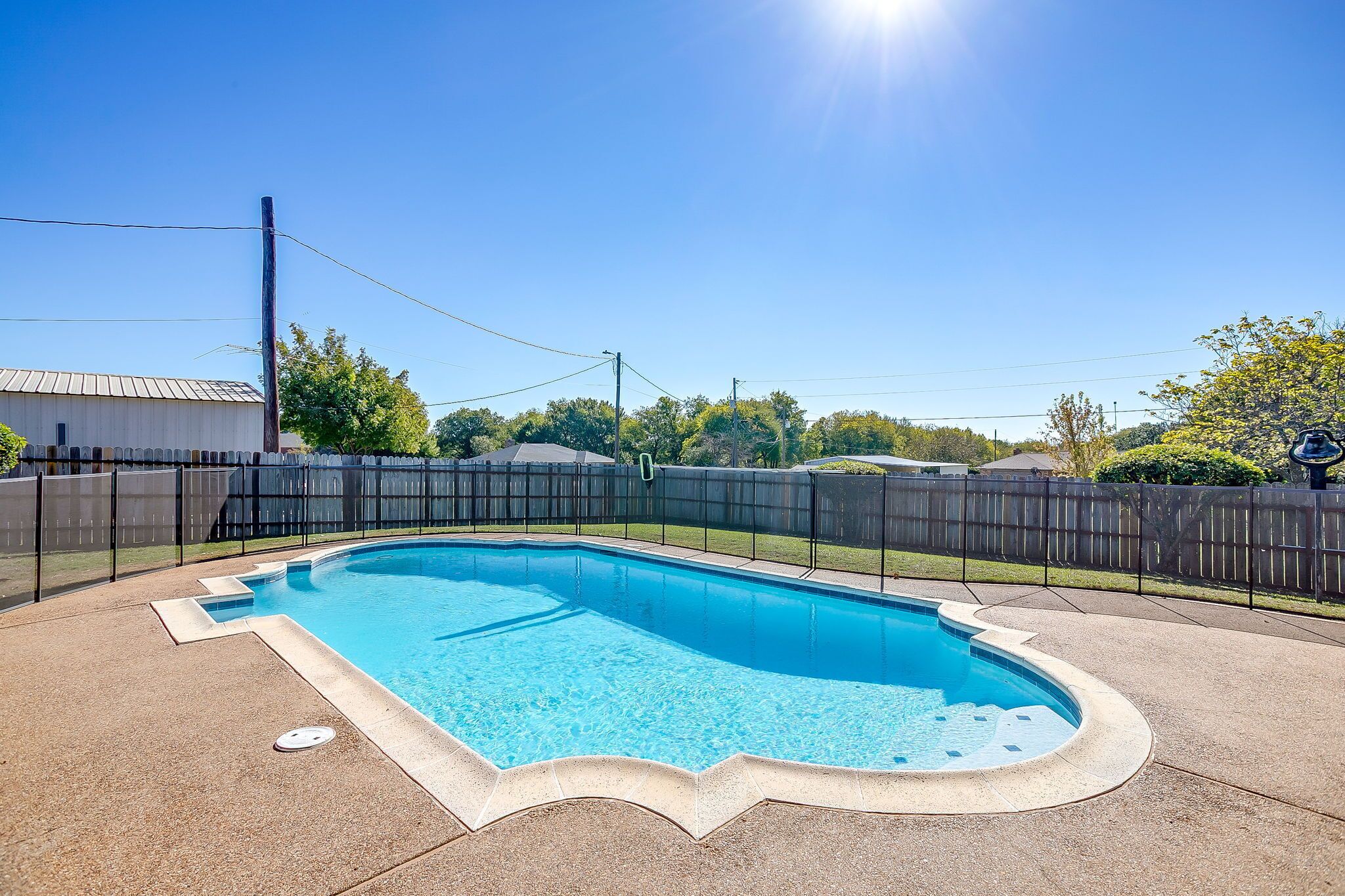 Une piscine rectangulaire à bord incurvé, entourée d’une terrasse en béton et d’une clôture noire.