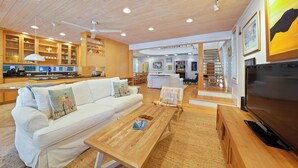 Ein modernes Wohnzimmer mit einem weißen Sofa, einem Holztisch und einem Fernseher. Es gibt eine Küchenzeile mit Holzschränken und einen Essbereich mit einem Stuhl.