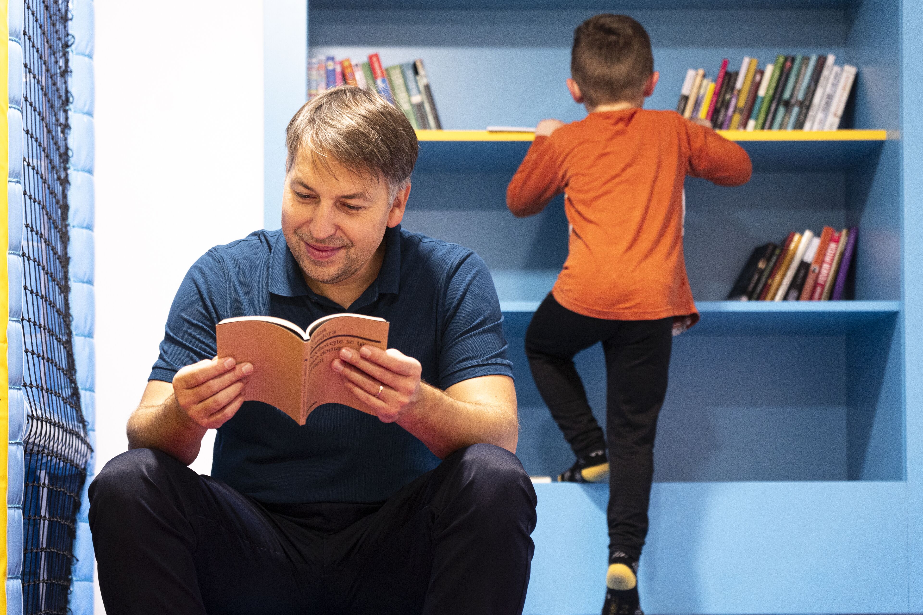 A man reading a book in a room with a bookshelf and a child in the background.