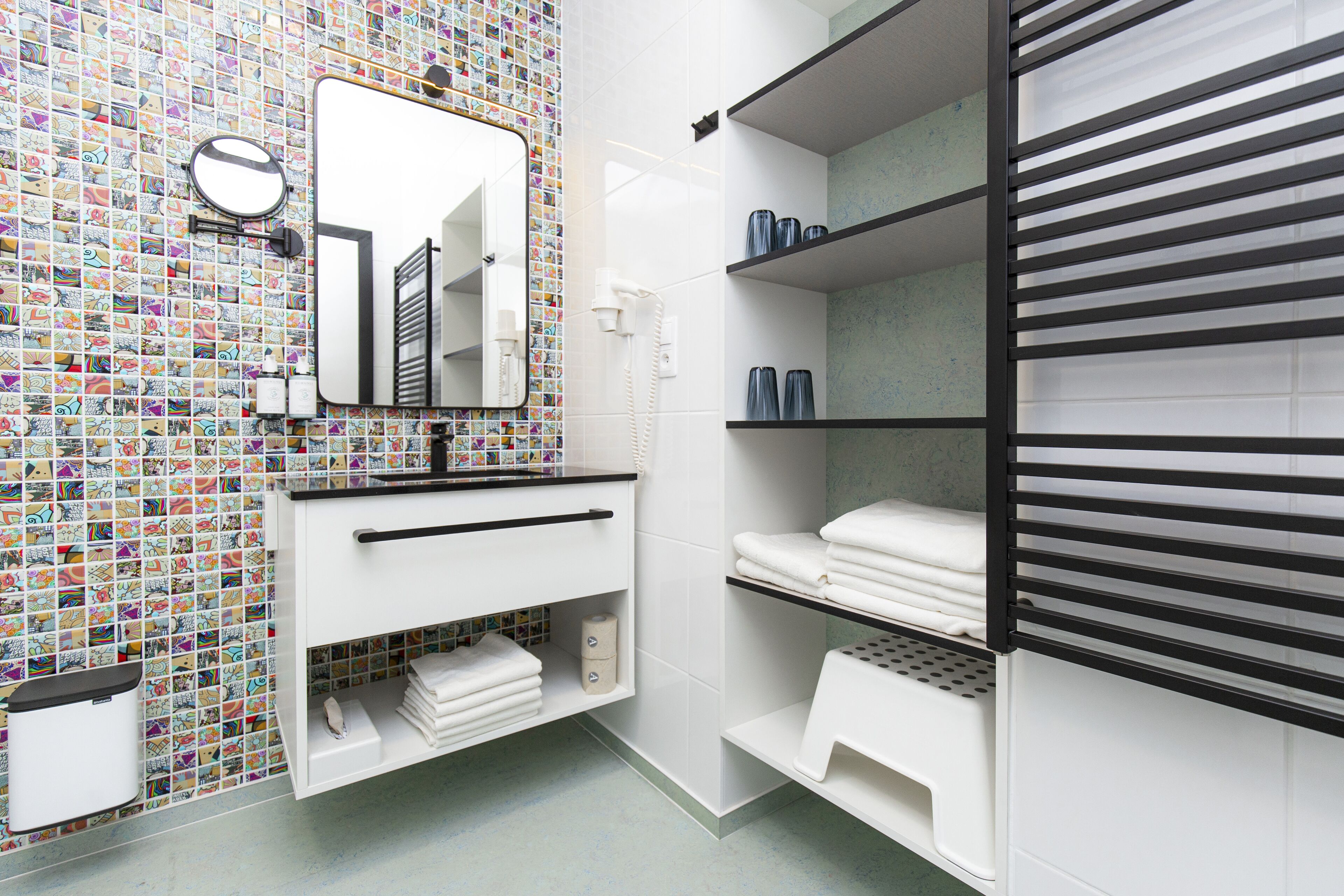 Un baño con pared de azulejos, espejo, lavamanos, estantes con toallas y un inodoro.