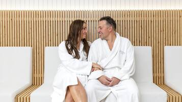 A couple in white bathrobes sitting on a white bench in a spa setting with wooden slats in the background.