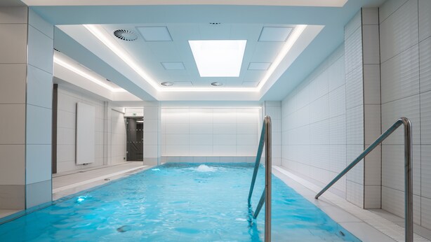An indoor swimming pool with clear blue water, a metal ladder, and a ceiling with recessed lighting.