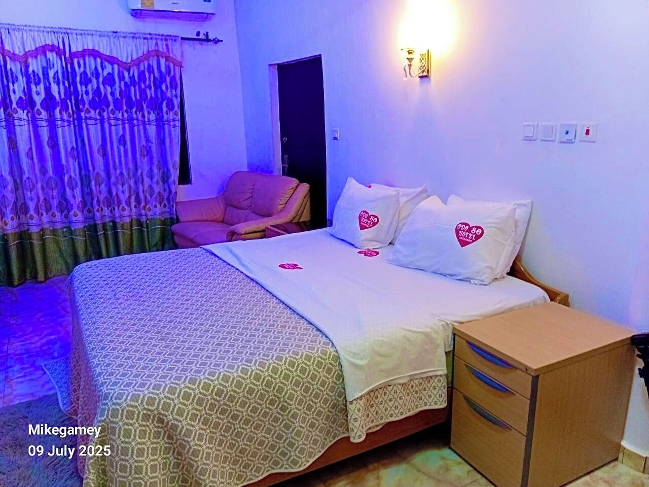 Un dormitorio con cama, una silla, una mesita de noche y una lámpara fijada a la pared.