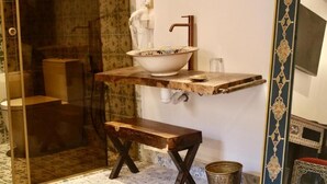 Un baño con cabina de ducha de vidrio, un banco de madera, un lavabo blanco con un cuenco decorativo y una pared con diseño.