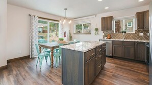 A modern kitchen with wooden cabinets, a granite countertop island, and a dining area with green chairs. - Hot tub! Dogs OK! King Bed & Fireplace, Near Park (Estes Park)
