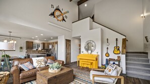 A spacious living room with a brown leather sofa, a wooden coffee table, and a patterned rug. A yellow piano stands in the center, and a staircase with a wooden railing leads to an upper level. - Hot tub! Dogs OK! King Bed & Fireplace, Near Park (Estes Park)