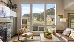 A cozy living room with a stone fireplace, large windows offering a view of houses and hills, a leather sofa, and a wooden coffee table. - Hot tub! Dogs OK! King Bed & Fireplace, Near Park (Estes Park)