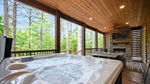 Un bain à remous avec une cheminée en pierre et un téléviseur fixé au mur, entouré d’une terrasse en bois et d’arbres.