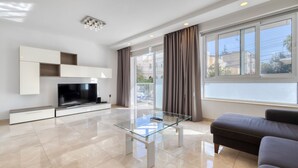 A modern living room with a flat-screen TV, a glass coffee table, a sofa, and a large window with curtains. - St Georges Bay Oasis, Steps from the Beach (Swieqi)