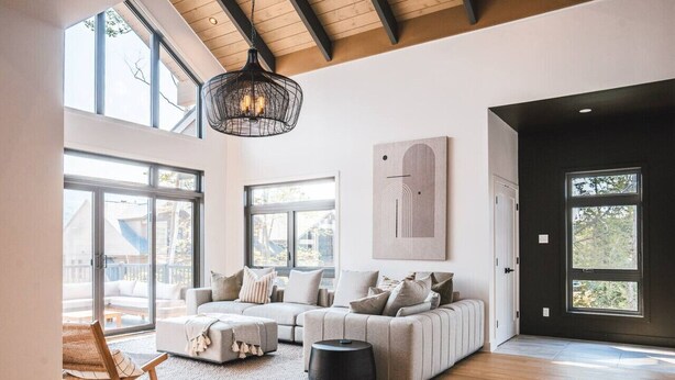 A spacious living room with a large sofa set, a coffee table, and a wicker chair. There is a black pendant light hanging from the ceiling, and a large window provides natural light.