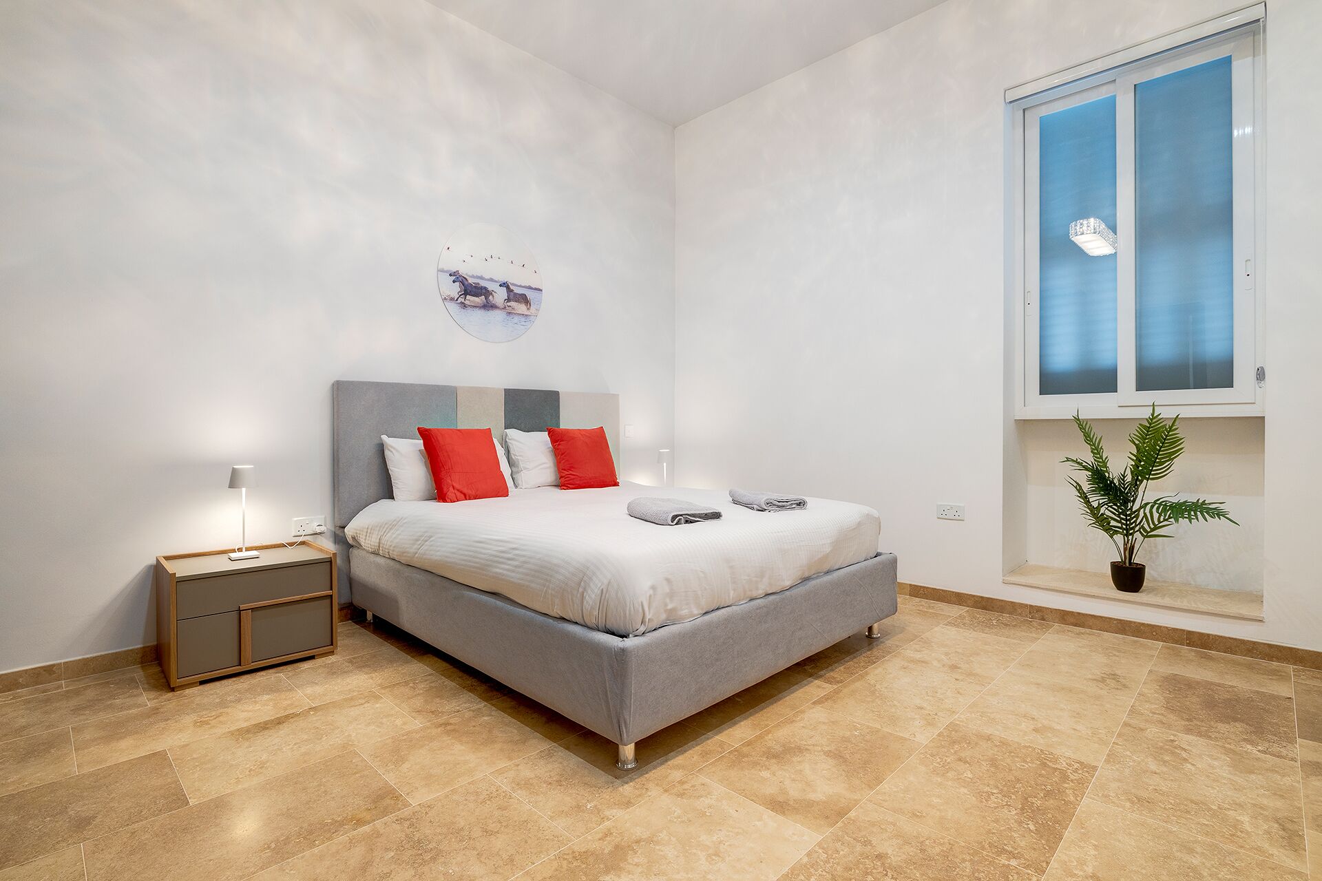 Un dormitorio con cama, mesita de noche, lámpara, ventana y planta en maceta.