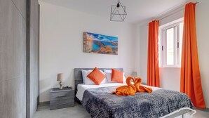 A modern bedroom with a bed, a nightstand, a painting, and orange curtains. - Modern Studio, Central Location (Gzira)