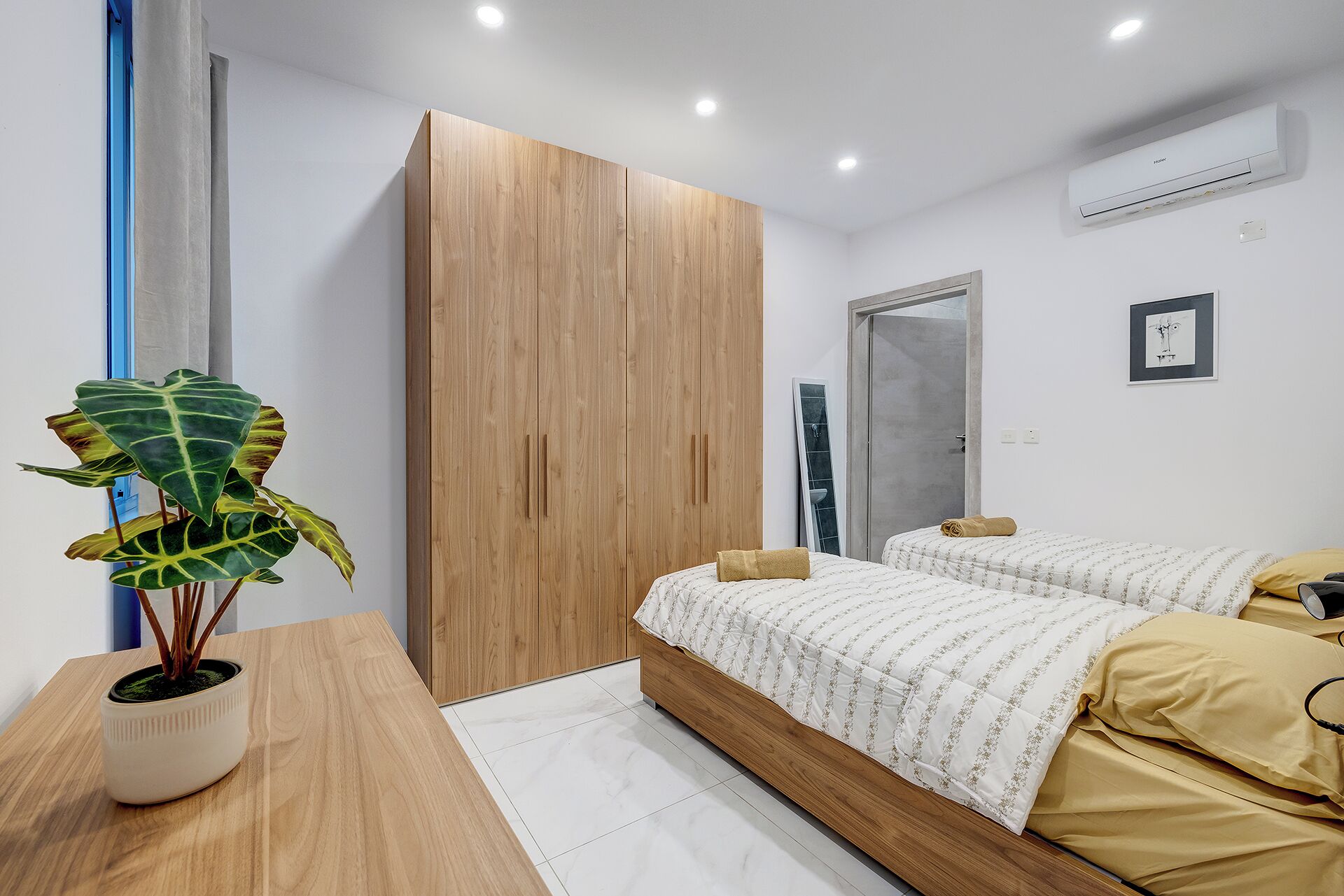 A modern bedroom with a wooden wardrobe, a bed with a patterned duvet, a bedside table with a plant, and an air conditioning unit on the wall.