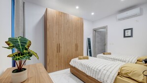 A modern bedroom with a wooden wardrobe, a bed with a patterned duvet, a bedside table with a plant, and an air conditioning unit on the wall. - Modern Family Holiday Retreat in Gzira (Gzira)